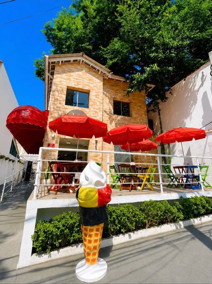 Fachada de tijolos da Gelateria Primo Amore com deck, guarda-sóis vermelhos e casquinha gigante na entrada